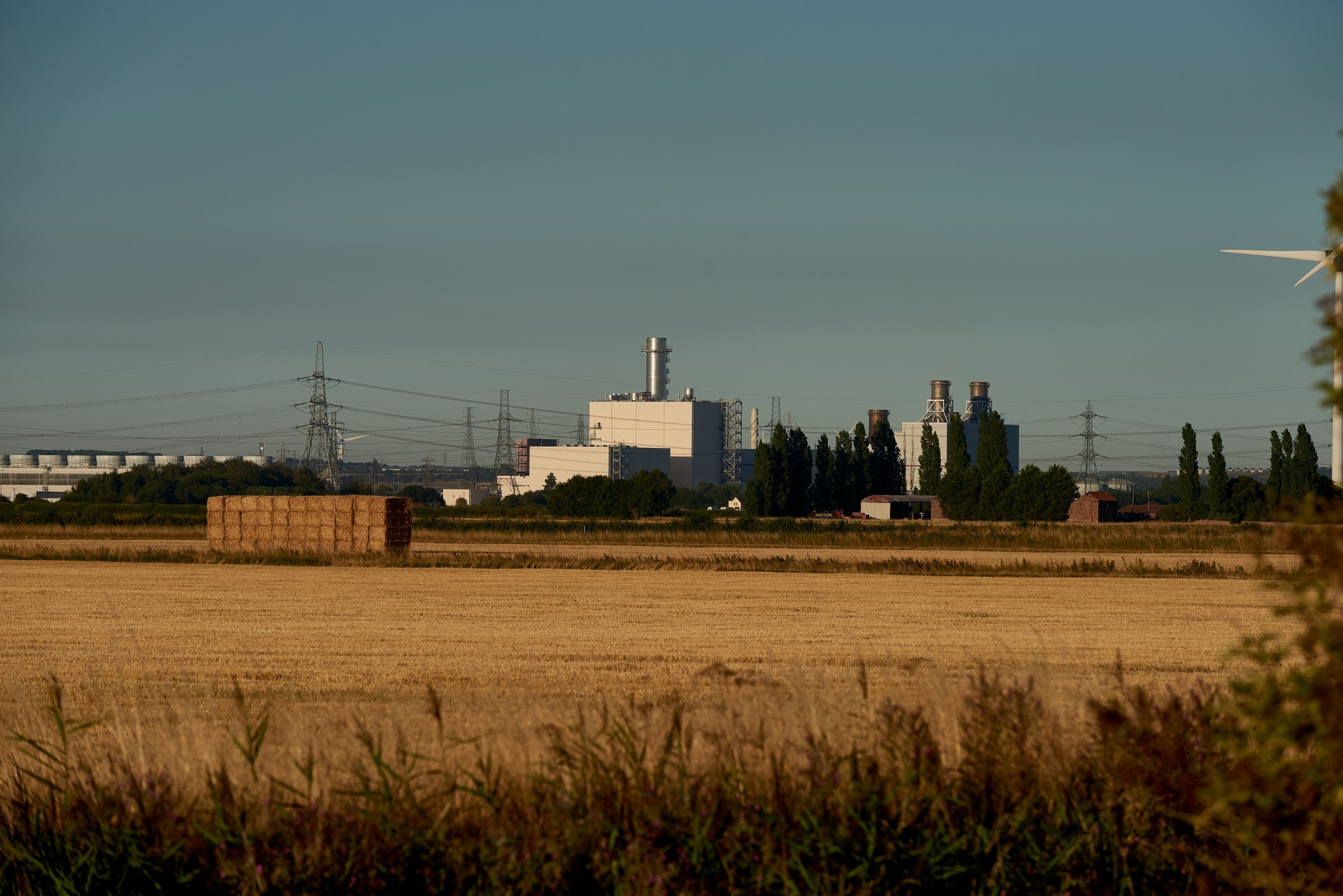 A large power station in the distance with fields and electricity pylons/towers and wires surrounding it